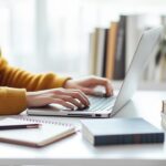 Modern ev ofisinde dizüstü bilgisayarda yazı yazan, not defteri ve kahveyle çevrili odaklanmış genç profesyonel.